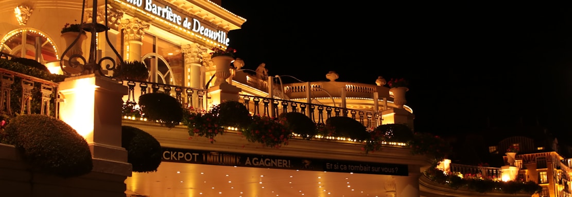 A luxurious casino at night, illuminated with warm, golden lights. The building features elegant architecture with columns and decorative elements. A few people are walking towards the entrance, and there are a couple of parked cars nearby.