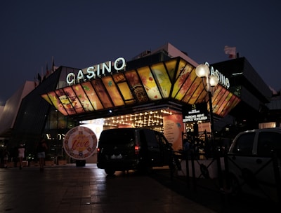 A building with a brightly illuminated entrance is seen at night. The sign above reads 'Casino' in bright letters. The facade features colorful graphics depicting various indulgent themes. Several vehicles are parked near the entrance, and people are visible in the vicinity. Tall lamp posts with spherical lights are present, casting a warm glow.